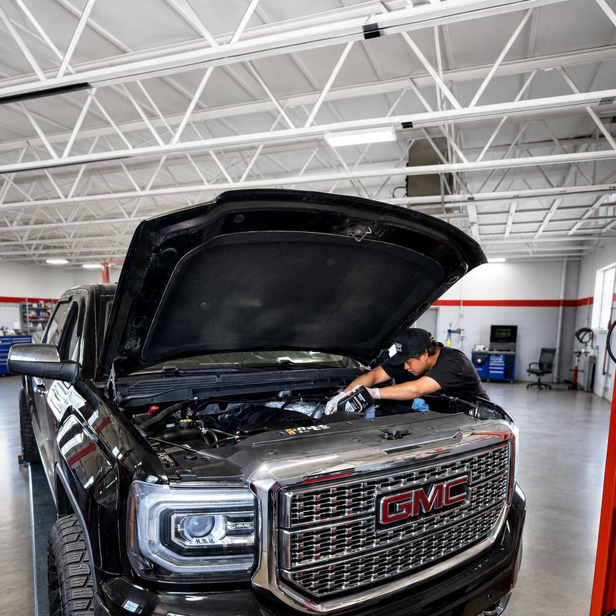 Technician working under the open hood of a black pickup truck in a service bay